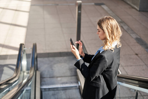 Porträt einer Frau auf der Rolltreppe, mit Handy und Blick auf den Smartphone-Bildschirm, die im schwarzen Anzug auf der Straße steht. - Foto, Bild