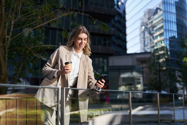 Successful businesswoman in suit, drinks her coffee and uses mobile phone, standing on street, having a lunch break. - Photo, Image