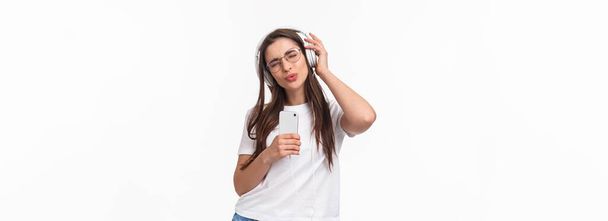 Technológia, életmód és zenei koncepció. Portré vidám, lelkes fiatal lány szemüvegben, dalok hallgatása fülhallgatóban, ének mentén félelmetes pálya, tartsa a mobiltelefon és a tánc. - Fotó, kép