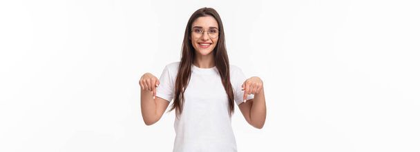 Klicken Sie hier. Porträt einer brünetten, wunderschönen jungen Frau mit Brille, zeigt mit dem Finger nach unten, um Werbung zu zeigen, lächelt zufrieden, schlägt Besuchslink vor, empfiehlt Download oder Abonnement, weißer Hintergrund. - Foto, Bild