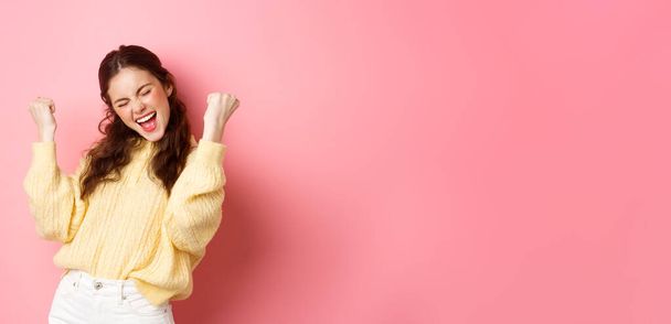 Girl screams with joy and fist pump, say yes, achieve goal or success, celebrating achievement, triumphing and winning, standing over pink background. - Photo, Image