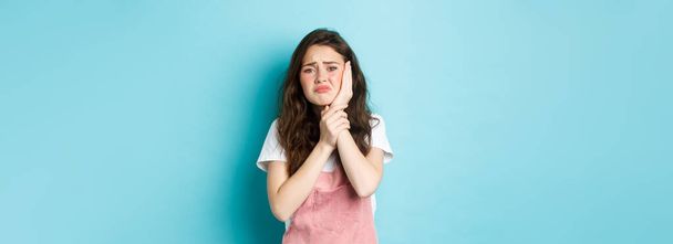 Image of frowning upset girl touching swallen cheek, having toothache, complaining on tooth decay, need to see dentist, standing against blue background. - Photo, image