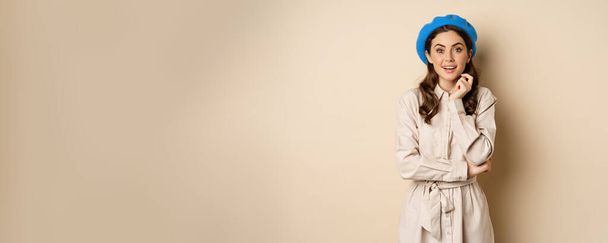 Image of stylsh beautiful woman looking surprised, shocked reaction at camera, posing in trench coat against beige background. - Фото, изображение