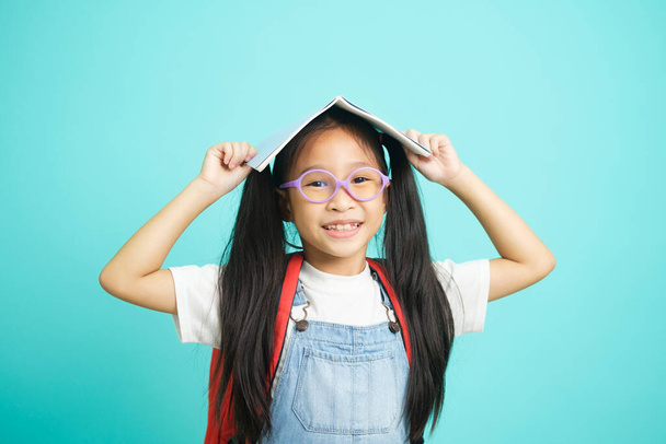 Portrait of happy lazy little girl covering head with book and smiling to camera, disobedient child having fun.indoor studio shot isolated on blue background.Educational concept for school - Photo, Image