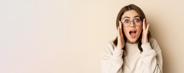 Close up portrait of cute woman in glasses looking impressed, reacting amazed at smth she sees, standing over beige background. - Foto, Imagem