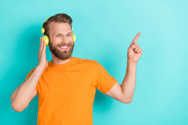 Photo of cheerful positive optimistic man dressed orange t-shirt indicating empty space touch headphones isolated on teal color background. - Photo, Image