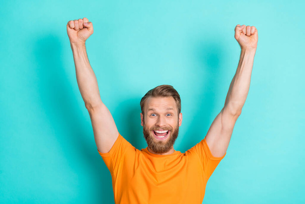 Photo of optimistic satisfied crazy man with blond hairstyle dressed orange t-shirt raise fists up isolated on teal color background. - Foto, Imagem