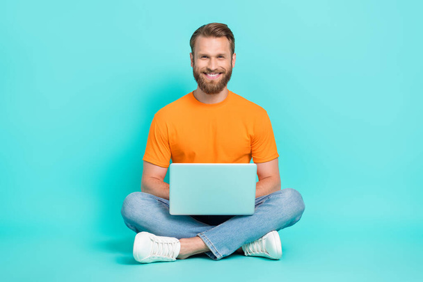 Full length photo of handsome guy with brown hair wear orange t-shirt sitting laptop on legs isolated on teal color background. - Fotoğraf, Görsel