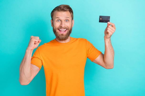 Photo of positive impressed man with masculine beard dressed orange t-shirt hold plastic credit card isolated on teal color background. - Foto, imagen