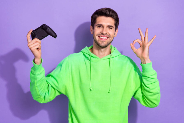 Photo of attractive satisfied guy with brunet hairdo dressed green hoodie hold joystick showing okey isolated on purple color background. - Foto, afbeelding