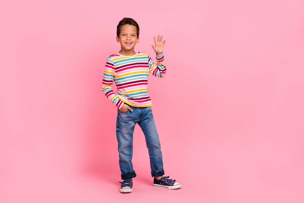 Full length photo of positive sweet optimistic boy dressed striped shirt jeans arm in pocket wave palm isolated on pink color background. - Zdjęcie, obraz