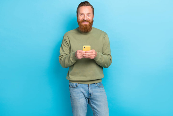 Photo of positive cheerful optimistic guy with red hairstyle dressed khaki long sleeve hold smartphone isolated on teal color background. - Fotó, kép