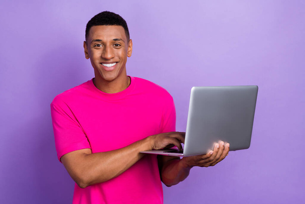 Photo of positive cheerful satisfied guy with box haircut dressed pink t-shirt typing on laptop isolated on purple color background. - Foto, Imagem
