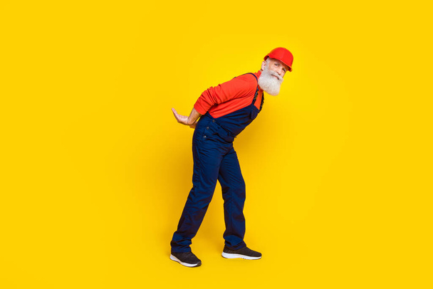 Photo of cute funny age man workwear overall red hard hat holding arms big empty space isolated yellow color background. - Foto, immagini