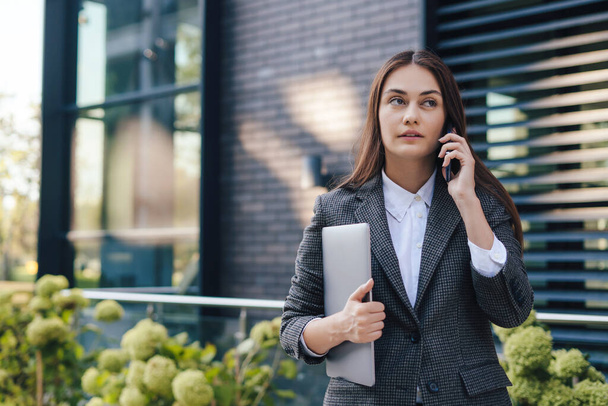 Brunette valkoihoinen liiketoiminnan nainen puhuu matkapuhelimella ulkona, kävely vastaan toimistorakennus. Kopioi tilaa. Käyttää kannettavaa tietokonetta ja matkapuhelinta. Etäisyys - Valokuva, kuva