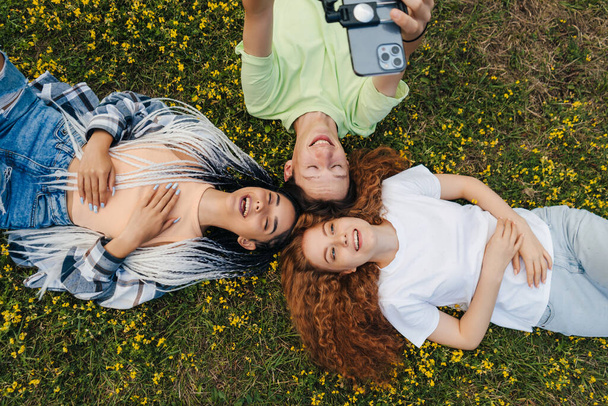 Tre giovani che si divertono insieme nel parco sdraiati sull'erba scattando selfie sul cellulare. Natura estiva. Stile di vita. Stile di vita felice. - Foto, immagini
