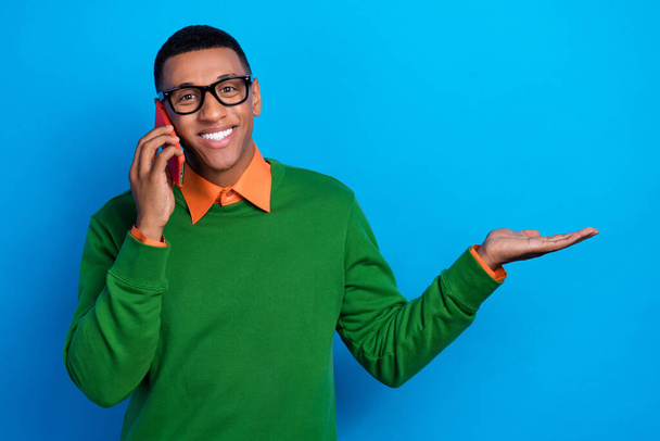 Photo of toothy beaming man orange shirt under green sweatshirt talk on phone demonstrate empty space isolated on blue color background. - 写真・画像