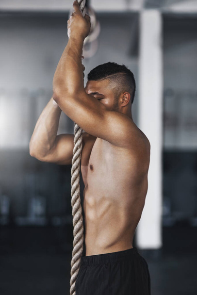 Η αναρρίχηση με σχοινί έχει να κάνει με τη δύναμη του σώματος. Ένας νεαρός άνδρας σκαρφαλώνει σε ένα σχοινί στο γυμναστήριο - Φωτογραφία, εικόνα
