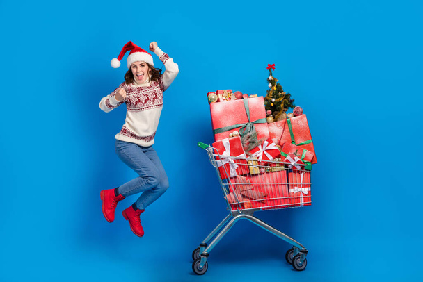 Photo of sweet lucky girl dressed print sweater claus cap running jumping choosing gifts supermarket tray isolated blue color background. - Фото, изображение