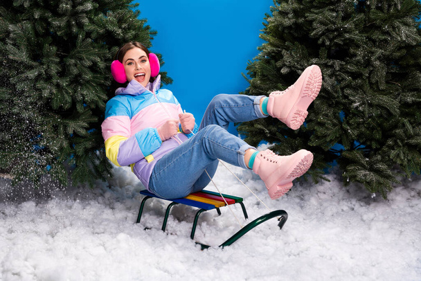 Photo of excited funky lady wear striped coat sledding between xmas fir indoors isolated blue color background. - Fotografie, Obrázek