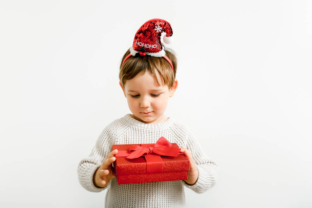 Portrait of little boy with Christmas headband holding red gift box on light wall background - Photo, Image