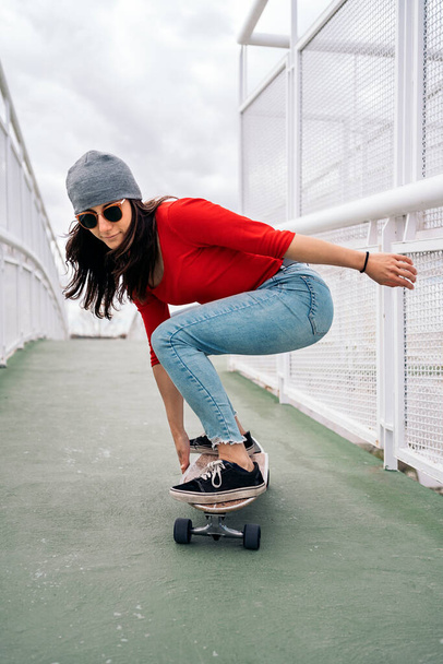 Cool skater woman practicing with her long-board in the city and having fun. She us wearing sunglasses and a hat. - Photo, Image
