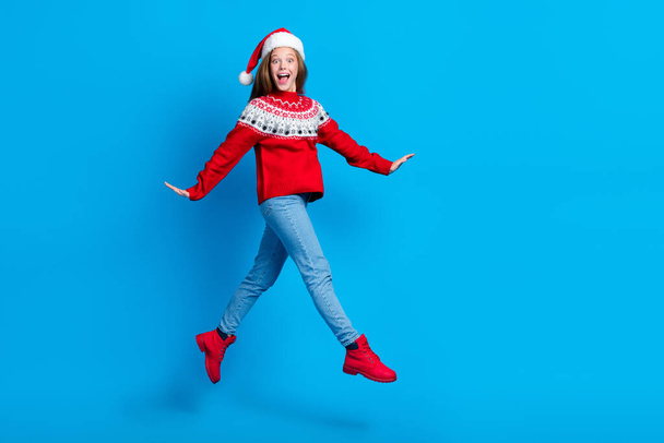 Volledige lengte foto van geschokt opgewonden school meisje dragen rode kerst ornament trui springen hoge lege ruimte geïsoleerde blauwe kleur achtergrond. - Foto, afbeelding