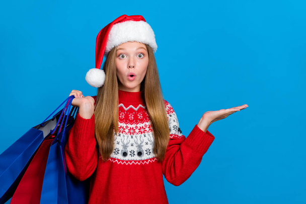 Portrait of impressed girl with blond hair wear red sweater hold shopping bags present empty space isolated on blue color background. - 写真・画像