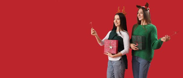 Noel havai fişekleri ve kırmızı arka planda mesajlaşmak için yeri olan hediyeleri olan güzel genç kadınlar. - Fotoğraf, Görsel