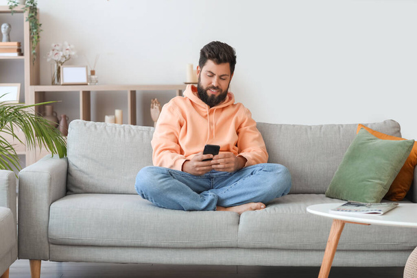 Красивий бородатий чоловік використовує мобільний телефон на сірому дивані вдома
 - Фото, зображення