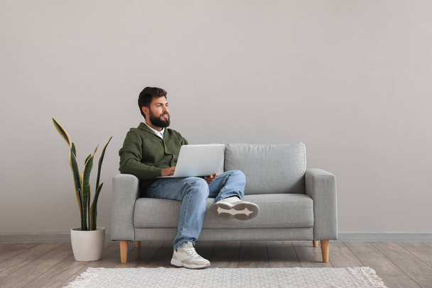 Schöner bärtiger Mann mit Laptop auf grauem Sofa nahe Lichtwand - Foto, Bild