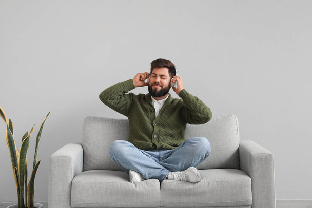 Handsome bearded man in headphones sitting on grey sofa near light wall - Fotoğraf, Görsel