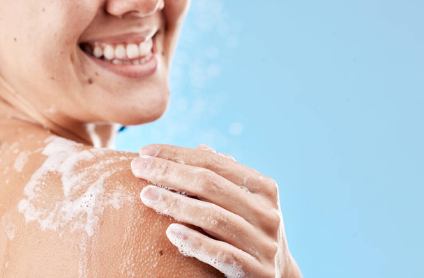 Shower, soap and smile of a woman grooming body for health, skincare and wellness against a blue mockup studio background. Water, cleaning and hand of a model with foam on shoulder and mock up space. - Foto, afbeelding