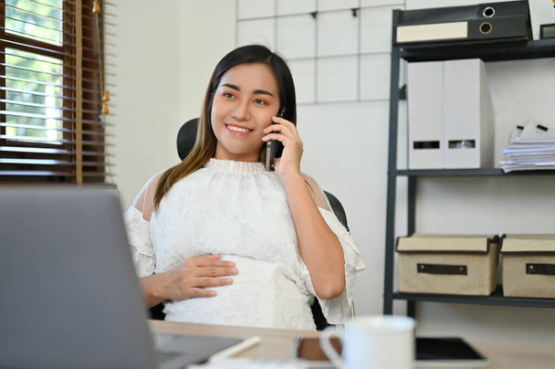Professionele en succesvolle Aziatische zwangere vrouwelijke baas of zakenvrouw praten aan de telefoon met haar zakelijke klant terwijl het aanraken van haar buik aan haar bureau. - Foto, afbeelding