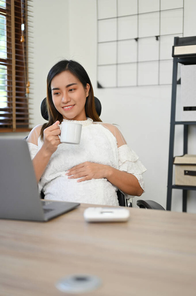 Portre, güzel ve profesyonel Asyalı hamile iş kadını ofisinde çalışırken çayını yudumluyor. İş ve hamilelik kavramı - Fotoğraf, Görsel
