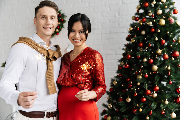 glücklicher Mann mit schwangerer asiatischer Frau mit glänzenden Wunderkerzen neben geschmücktem Weihnachtsbaum zu Hause - Foto, Bild