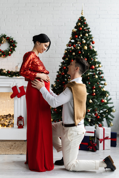 Diz çöküp hamile ve zarif Asyalı karısına Noel ağacının yanında sarılan adamın yan görüntüsü.  - Fotoğraf, Görsel