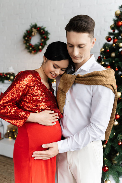 mężczyzna dotykając brzuch uśmiechnięty i w ciąży Azji żona w uroczyste ubrania w pobliżu rozmyte świąteczne wystrój - Zdjęcie, obraz