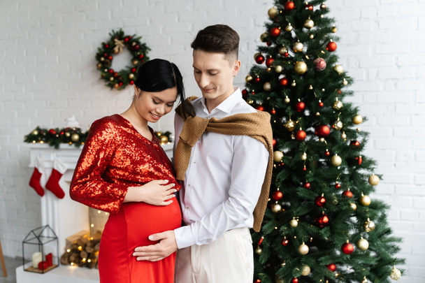 νεαρός άνδρας αγγίζοντας την κοιλιά της εγκύου Ασιάτισσας συζύγου σε εορταστικά ρούχα στο σαλόνι με χριστουγεννιάτικη διακόσμηση - Φωτογραφία, εικόνα