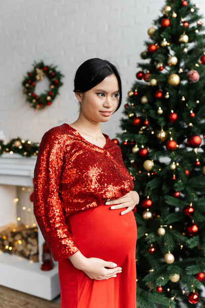 embarazada mujer asiática en rojo y brillante blusa tocando la barriga y mirando hacia otro lado en sala de estar con decoración de Navidad - Foto, Imagen