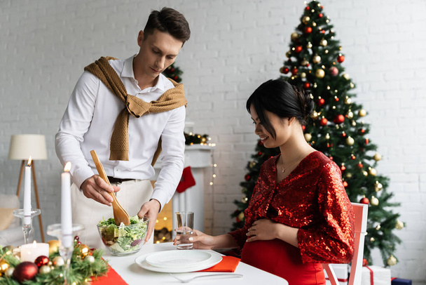 zwanger aziatische vrouw glimlachen in de buurt man met verse groente salade tijdens romantisch kerstdiner thuis - Foto, afbeelding