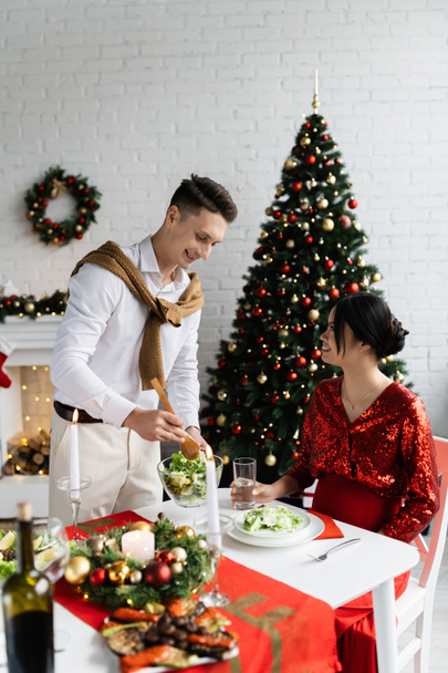 happy man holding bowl of vegetable salad near pregnant asian woman during Christmas supper at home - Valokuva, kuva