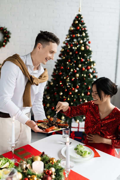 smiling man holding plate with grilled vegetables near asian wife with fork during romantic Christmas supper - Foto, imagen