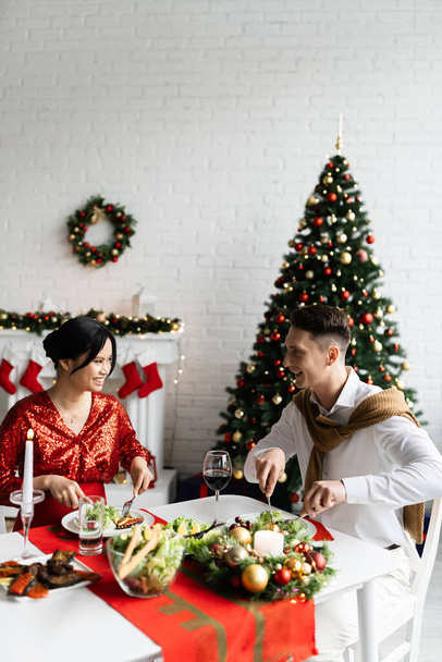 Ευτυχισμένος άντρας και η έγκυος Ασιάτισσα γυναίκα του να χαμογελούν ο ένας στον άλλο ενώ τρώνε Χριστουγεννιάτικο δείπνο στο σπίτι. - Φωτογραφία, εικόνα