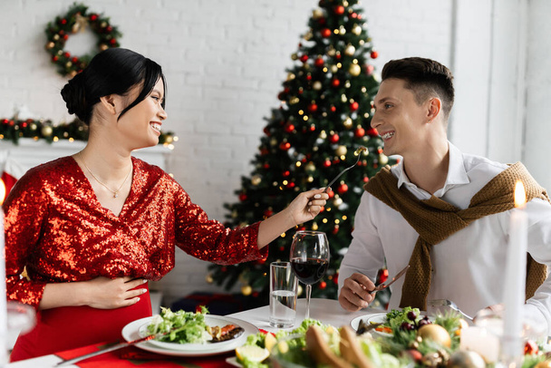 cheerful and pregnant asian woman feeding husband while having fun during romantic Christmas supper  - Foto, Imagen