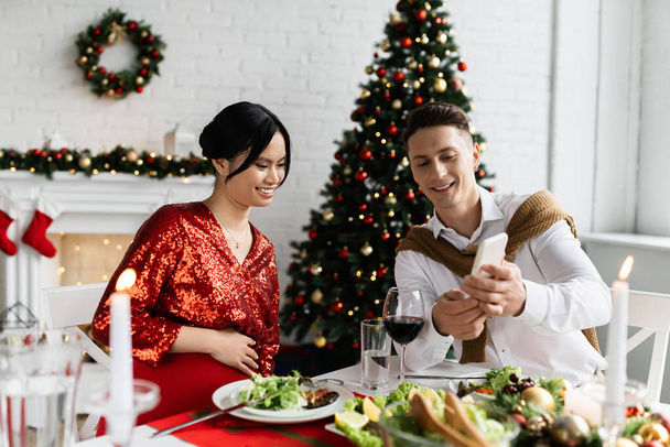 улыбающийся мужчина показывает смартфон беременной азиатской жене во время романтического рождественского ужина дома - Фото, изображение