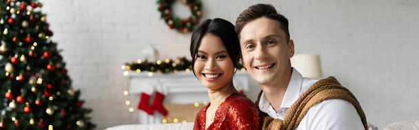 νεαρό πολυεθνικό ζευγάρι χαμογελά στην κάμερα κοντά θολή διακόσμηση Χριστουγέννων στο σπίτι, πανό - Φωτογραφία, εικόνα