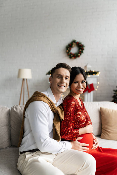 cheerful man with pregnant asian wife in festive outfit looking at camera on couch at home - Foto, Imagen
