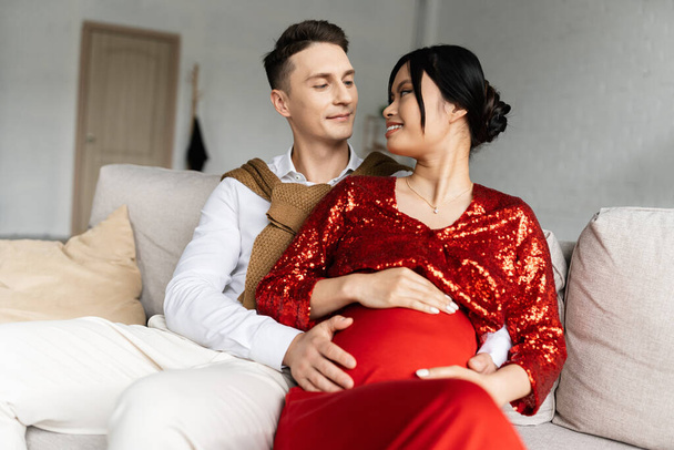 pregnant asian woman in red and shiny clothes smiling at young husband hugging her tummy on couch at home - Foto, Bild