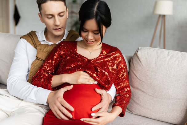 young man embracing belly of pregnant asian wife wearing red and shiny clothes  - 写真・画像